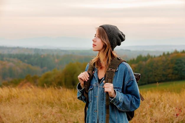 Style woman in denim jacket and hat with backpack in countryside with mountains