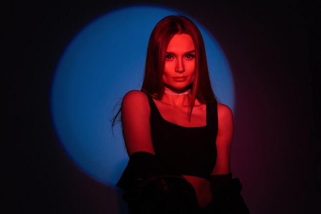 Styling beautiful young girl in fashionable black rock clothes with a leather jacket and a top in red light color on a blue circular light in the studio on a dark background Colorful portrait