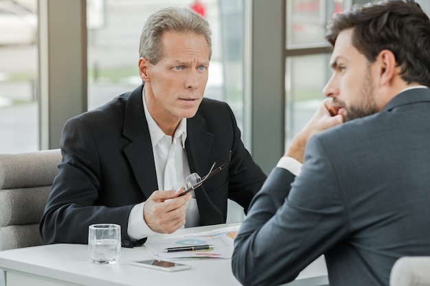 Stylish adult businessmen while working day in office. Businessmen seriously discussing plan. Office interior with big window