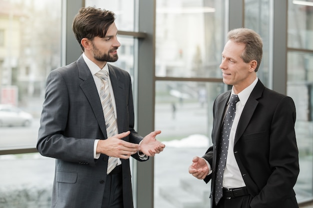 Stylish adult businessmen while working day in office. Businessmen talking. Office interior with big window