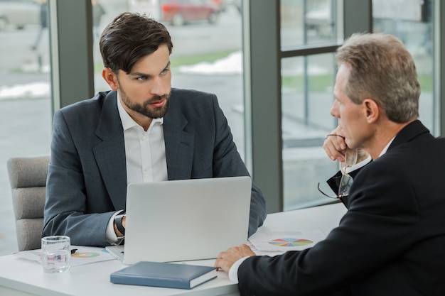 Stylish adult businessmen while working day in office. Businessmen working with laptop. Office interior with big window