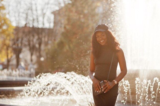 Stylish african american girl in gray tunic crossbody bag and cap posed at sunny autumn day against fountains Africa model woman