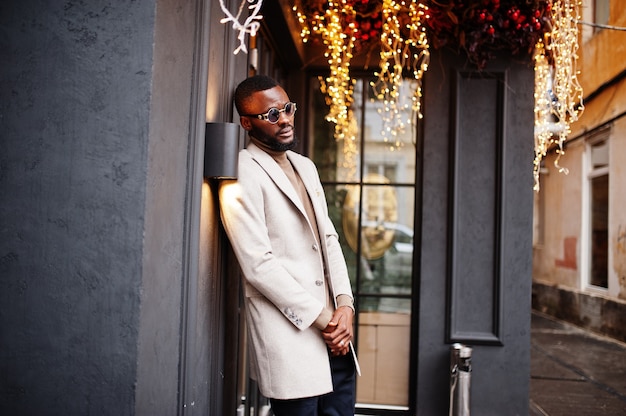 Stylish african american man wear beige jacket with sunglasses pose against house with golden garland.