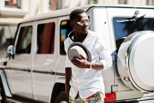 Stylish african american man in white shirt and colored pants with hat and glasses posed outdoor against silver mafia suv car Black fashionable model boy