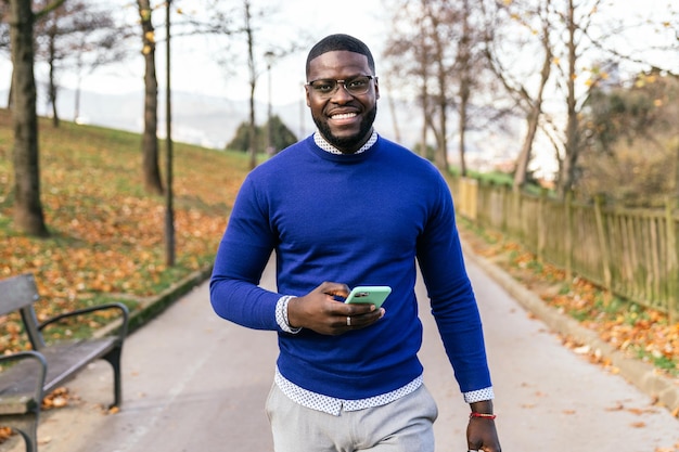 Stylish African Youth Radiates Joy Sporting Blue Sweater and Glasses Captured with Mobile in Hand in Beautifully Lit Park