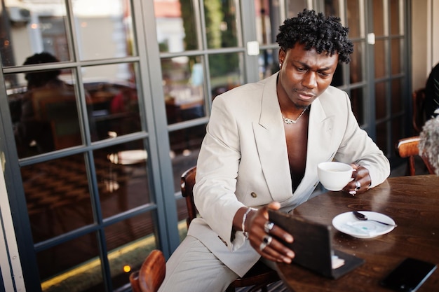 Stylish afro man in beige old school suit sitting on cafe with cup of coffee Fashionable young African male in casual jacket on bare torso