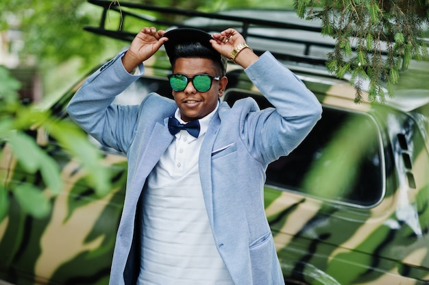 Stylish arabian man at sunglasses, cap and bow tie posed against military car. Arab model man.