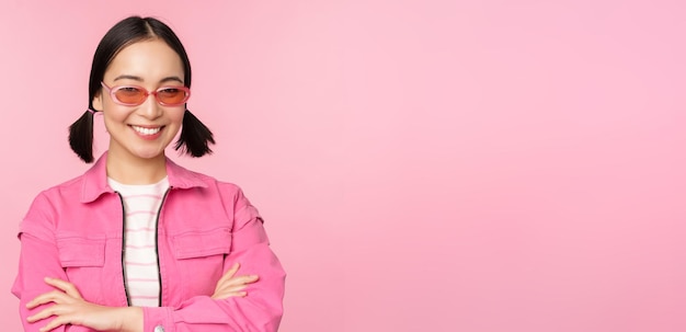 Stylish asian girl in pink clothes and sunglasses smiling and looking happy at camera standing over