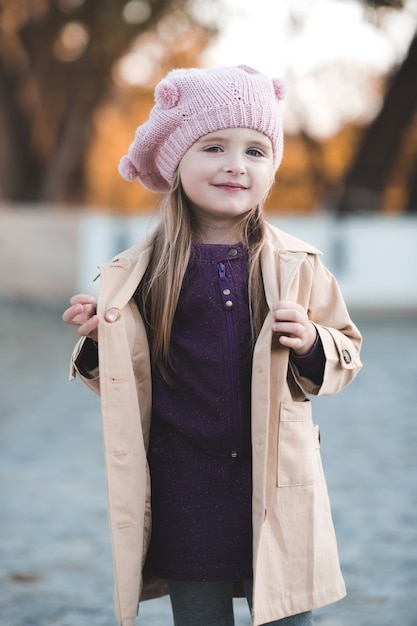 Photo stylish baby girl wearing trendy coat and knitted hat walking in park