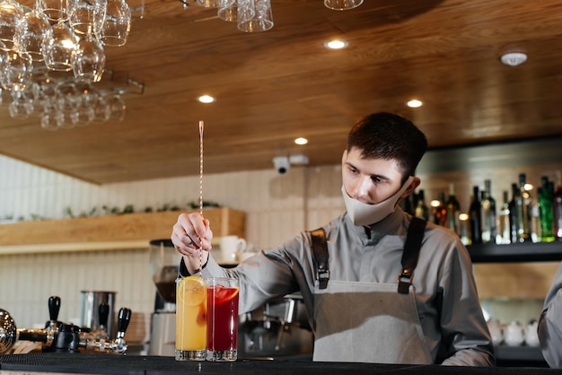 A stylish bartender in a mask and uniform during a pandemic is preparing cocktails at a party. The work of restaurants and cafes during the pandemic.