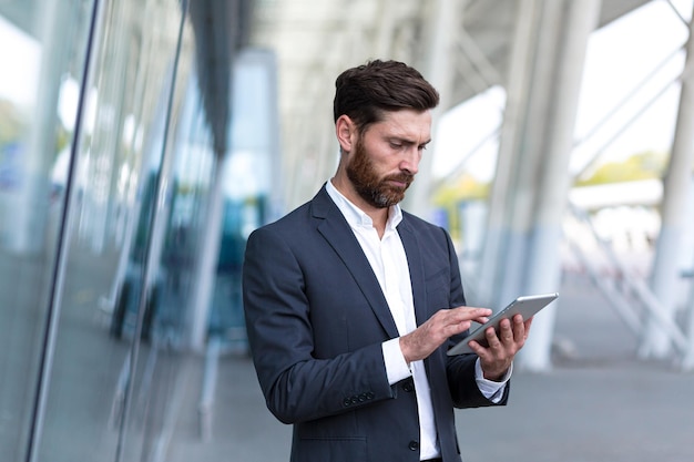 Photo stylish bearded businessman in formal business suit standing working with tablet in hands on background modern office building outside. man using smartphone or uses mobile phone outdoors city street