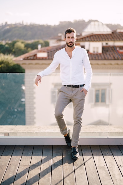 Stylish bearded guy in a white shirt and light trousers on a rooftop terrace in Florence, Italy