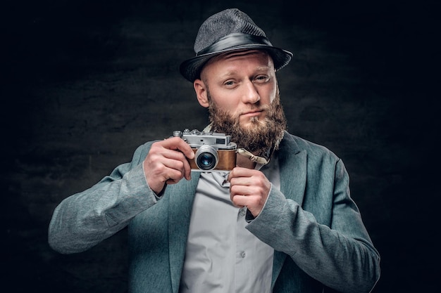 A stylish bearded hipster male dressed in a grey jacket and felt hat holds an SLR photo camera.