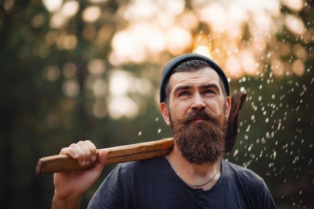 Stylish bearded man lumberjack with a long beard and mustache in a T-shirt and a warm hat with an ax on his shoulder