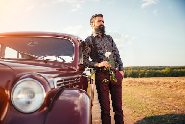 Stylish bearded man with a mustache in classic trousers with suspenders and a dark shirt with a rose in his hands at a brown retro car