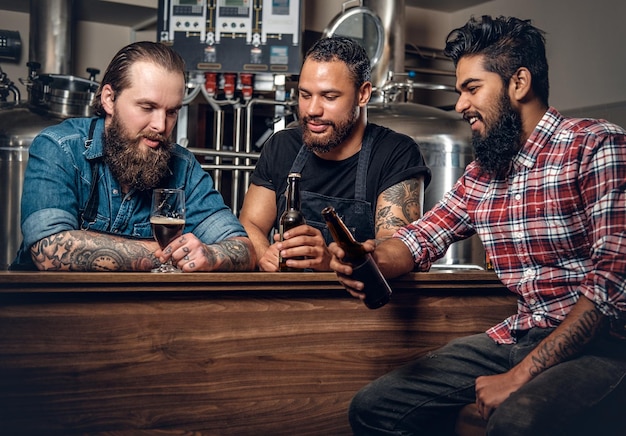 Stylish bearded tattooed Caucasian, Black and Indian men drinking craft beer in the microbrewery.