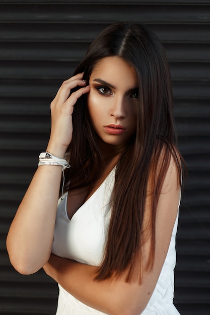 Stylish beautiful american woman in a white dress near a dark wall