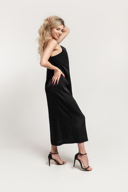 Stylish beautiful blonde woman in a fashion black elegant dress with high heels shoes posing in the studio on a white background Happy party girl with slender sexy body