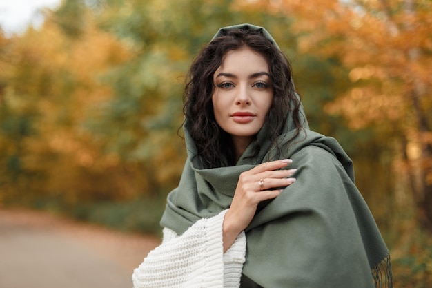 Stylish beautiful fresh woman with a curly hairstyle with a fashion green scarf on her head in a knitted vintage sweater walks in the autumn park