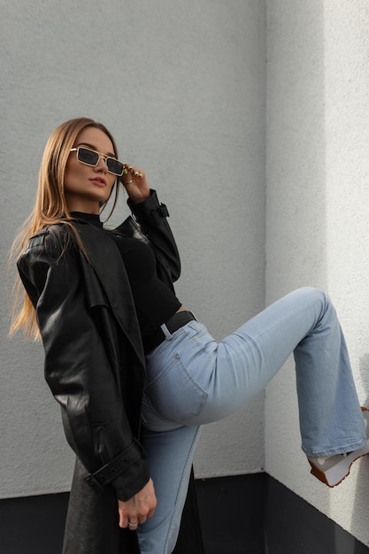 Stylish beautiful model girl hipster in fashionable clothes with a long leather coat and vintage blue high waisted jeans adjusts sunglasses and poses near a wall on the street