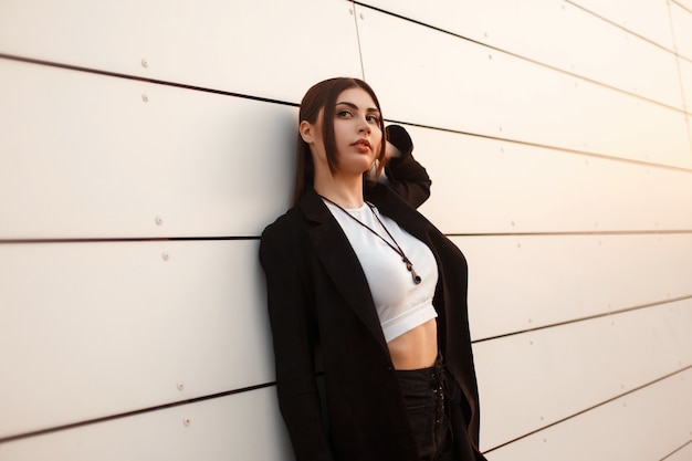 Stylish beautiful woman in a black fashion coat posing near a white wall