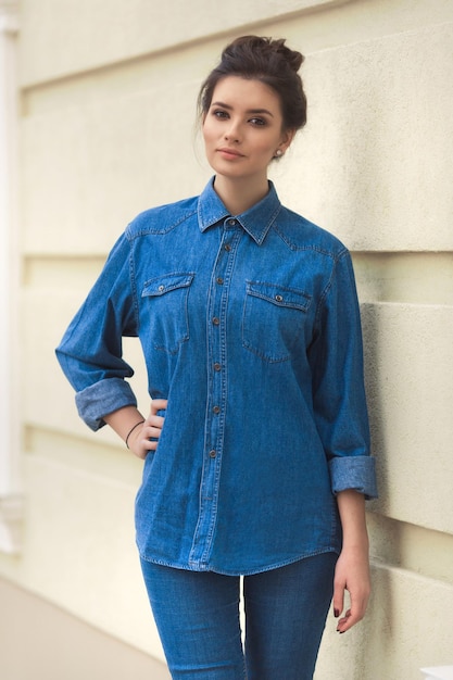 Stylish beautiful woman in denim shirt set standing near the wall Street style fashion total denim