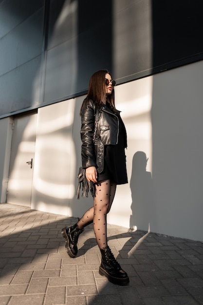 Stylish beautiful woman in fashionable black clothes with a stylish bag wearing sun goggles walks near the building in the city at sun light