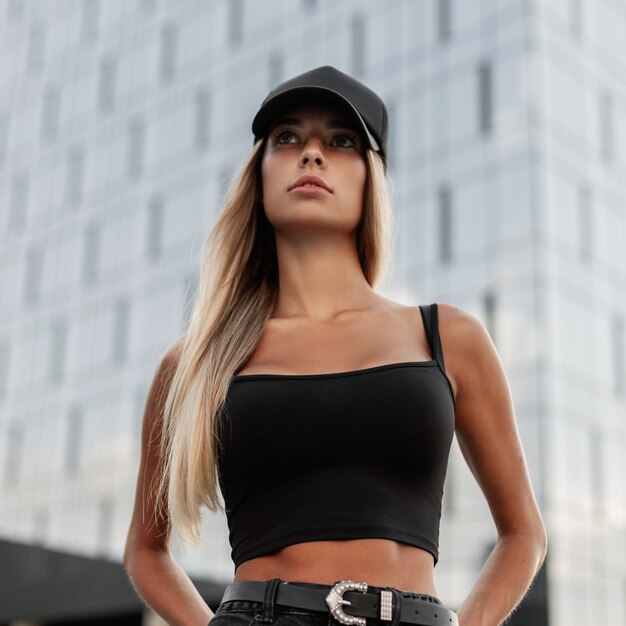 Stylish beautiful woman with a black fashionable cap with a mockup for design in a black t-shirt stands in the city