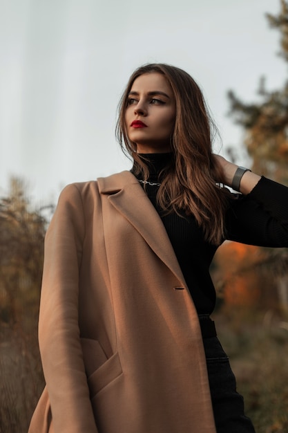 Stylish beautiful woman with red lips in fashionable clothes with a coat and a sweater stands in nature