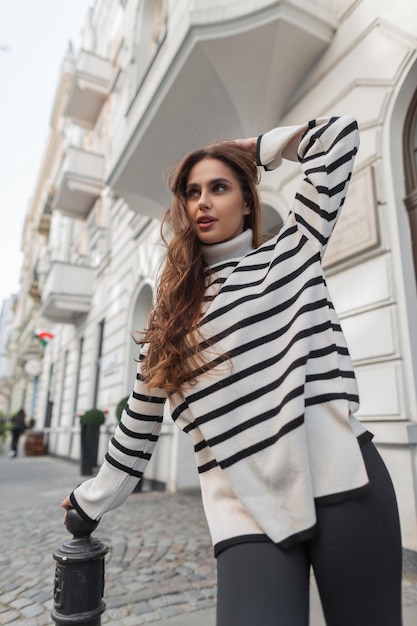 Stylish beautiful young brunette girl in a trendy fashion striped sweater walks and poses in the city