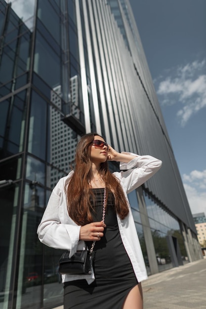 Stylish beautiful young chic lady with sunglasses in a black trendy fashion dress with a white shirt with a handbag walks in a sunny modern city