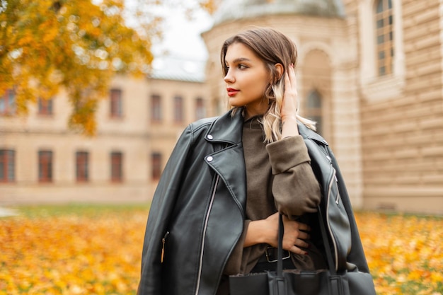 Stylish beautiful young girl in fashionable casual clothes with a black leather jacket a green sweater and a bag walks near the vintage city in the autumn city with yellow leaves