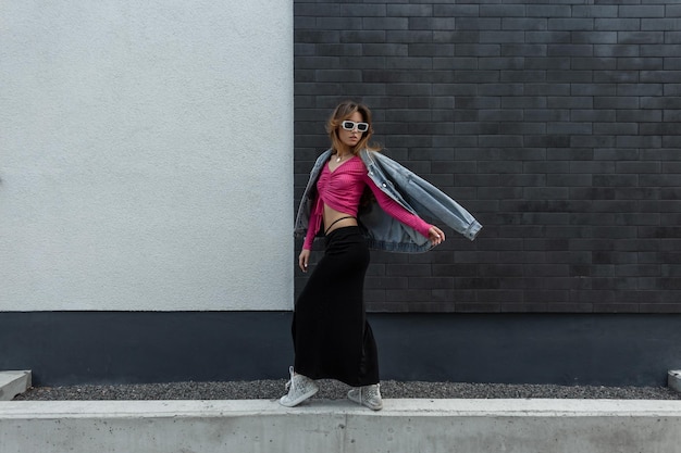 Stylish beautiful young woman model in fashionable street clothes with a pink crop top jeans jacket and a long skirt with sneakers walking near a brick wall in the city Urban female style look