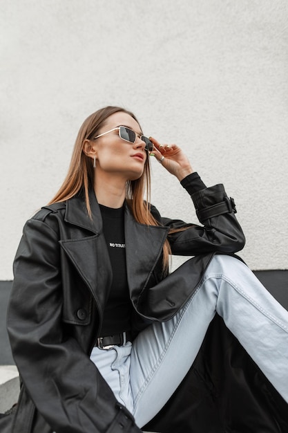 Stylish beautiful young woman model wears a sunglasses in fashionable casual urban clothes sits on the street near a white wall