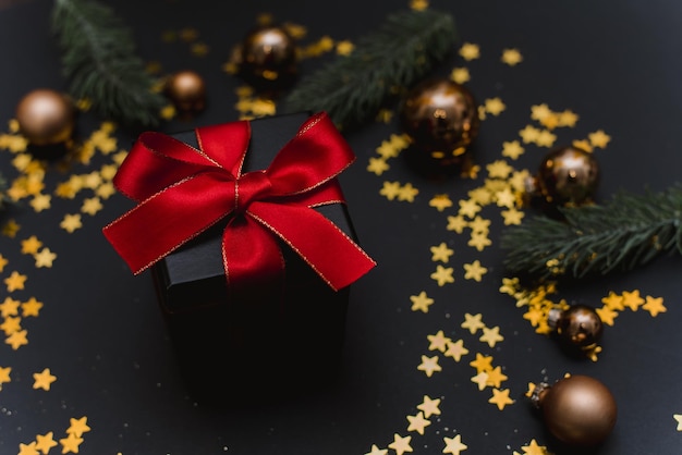 Stylish black gift with a red bow on the background of Christmas decorations confetti and fir branches