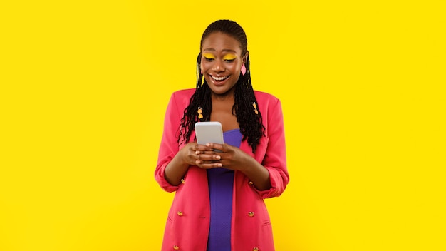 Stylish black lady using phone surfing apps over yellow background