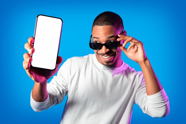 Stylish black man wearing sunglasses showing smartphone with empty screen