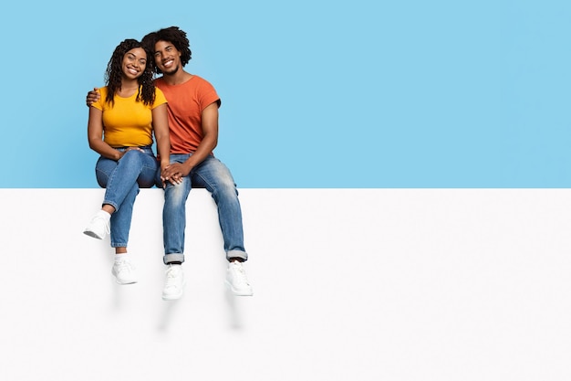 Stylish black people sitting on empty advertising board hugging