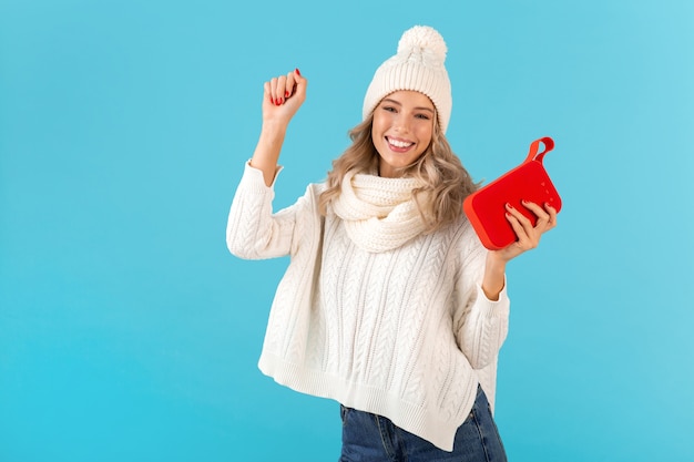 Stylish blond smiling beautiful young woman holding wireless speaker listening to music happy dancing wearing white sweater and knitted hat winter style fashion posing 