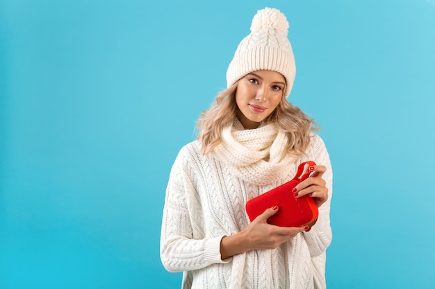 Stylish blond smiling beautiful young woman holding wireless speaker listening to music happy wearing white sweater and knitted hat winter style fashion posing isolated on blue wall