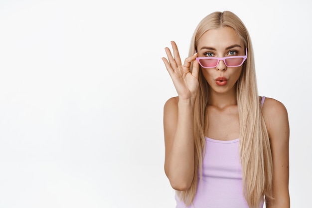 Stylish blond woman looking surprised, take-off sunglasses and stare amazed, standing in tank-top on white