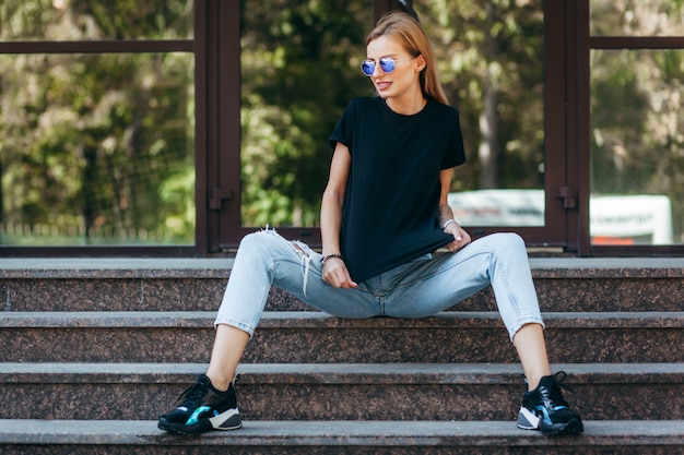 Stylish blonde girl wearing black t-shirt and glasses posing against street