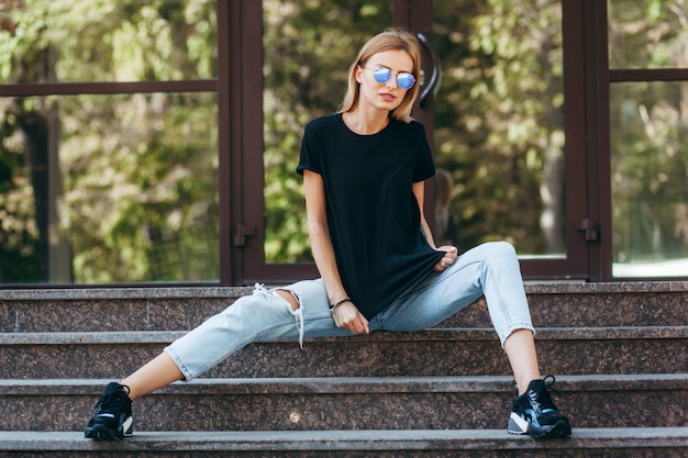 Stylish blonde girl wearing black t-shirt and glasses posing against street