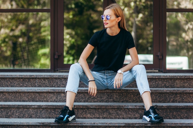 Stylish blonde girl wearing black t-shirt and glasses posing against street