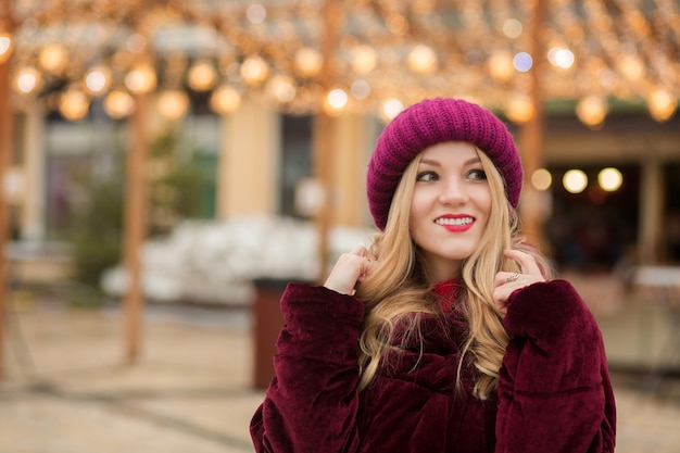 Stylish blonde woman wearing red knitted hat and winter coat, posing on the background of garland at the street in Kyiv