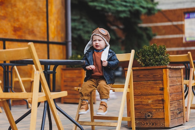 Stylish boy 3 years old in a leather jacket and brown trousers sits on the terrace near the cafe Modern child Children's fashion Happy child