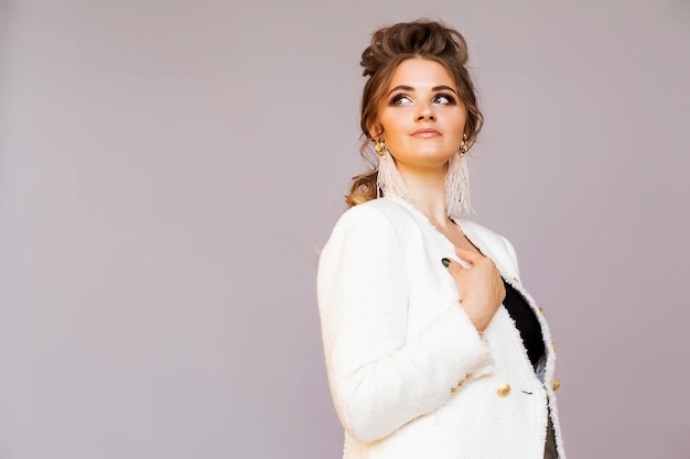 Stylish brunette girl posing on a gray background in a stylish white jacket with ostrich feather earrings