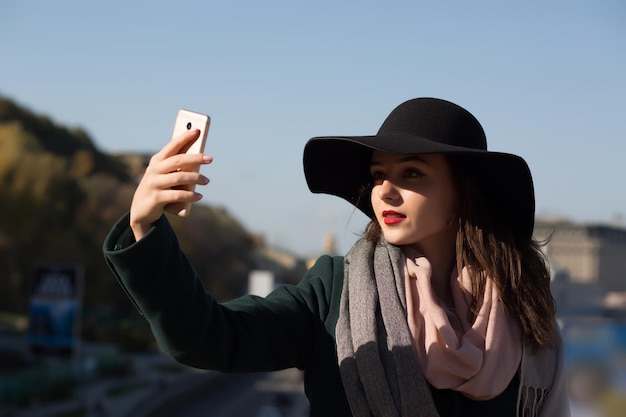 Stylish brunette girl tourist in hat taking a selfie photo in sunny day in Kiev