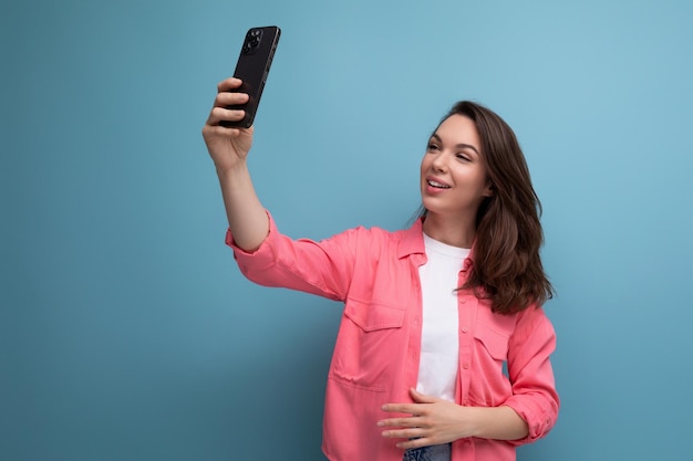 Stylish brunette woman in a trendy shirt takes a selfie using a phone on a studio background