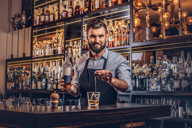 Stylish brutal bartender in a shirt and apron makes a cocktail at bar counter background.